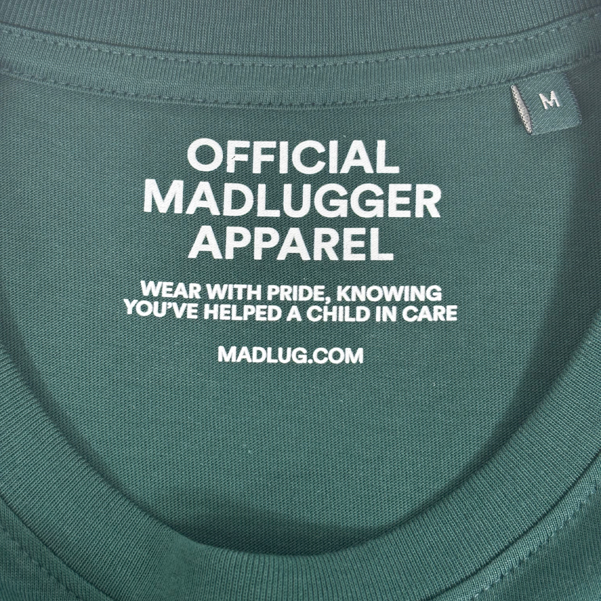 Close-up of a dark green t-shirt neck label with white printed text reading 'OFFICIAL MADLUGGER APPAREL' and 'WEAR WITH PRIDE, KNOWING YOU'VE HELPED A CHILD IN CARE' along with 'MADLUG.COM'