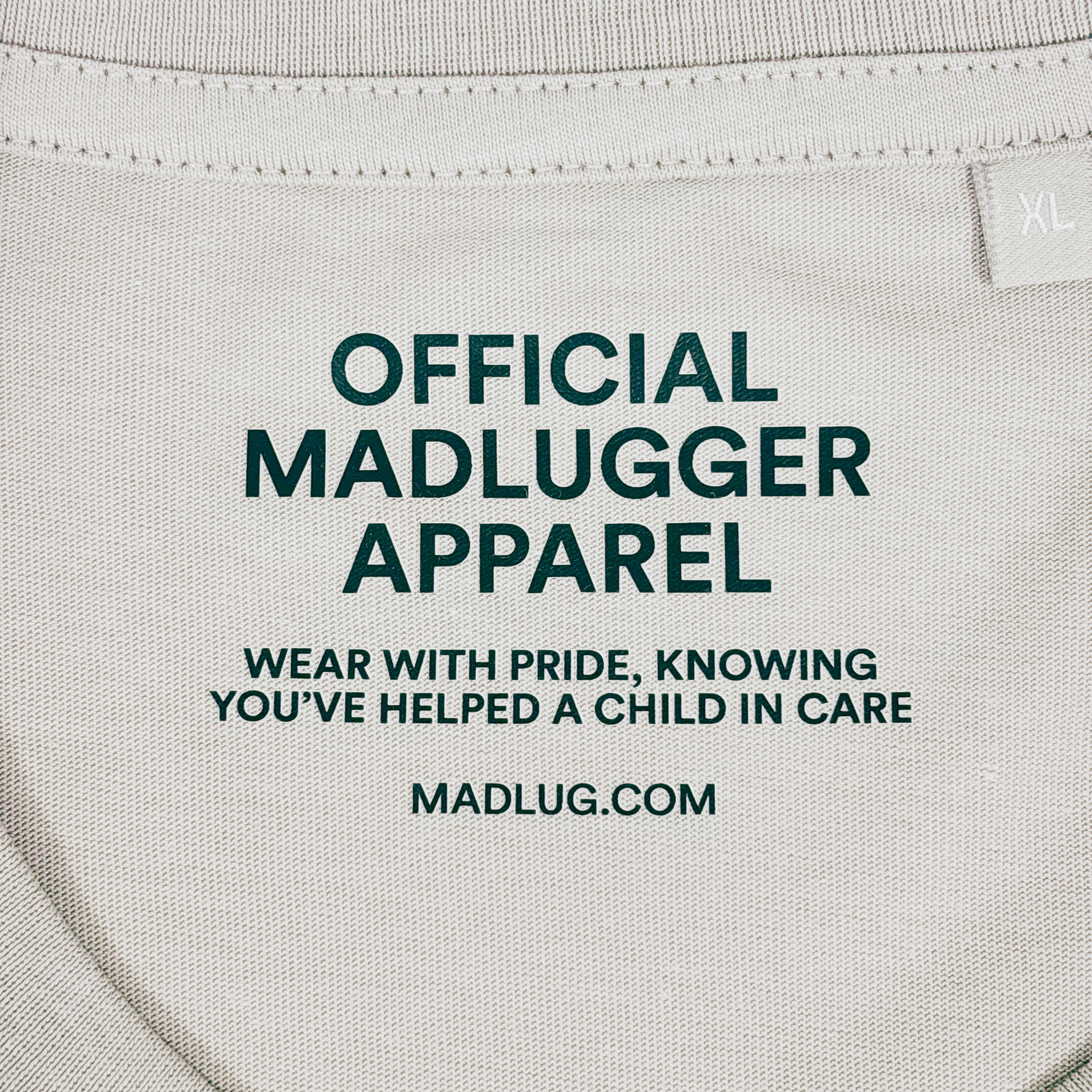 Close-up of a stone colour t-shirt neck label with black printed text reading 'OFFICIAL MADLUGGER APPAREL' and 'WEAR WITH PRIDE, KNOWING YOU'VE HELPED A CHILD IN CARE' along with 'MADLUG.COM'