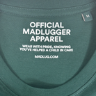 Close-up of a dark green t-shirt neck label with white printed text reading 'OFFICIAL MADLUGGER APPAREL' and 'WEAR WITH PRIDE, KNOWING YOU'VE HELPED A CHILD IN CARE' along with 'MADLUG.COM' 