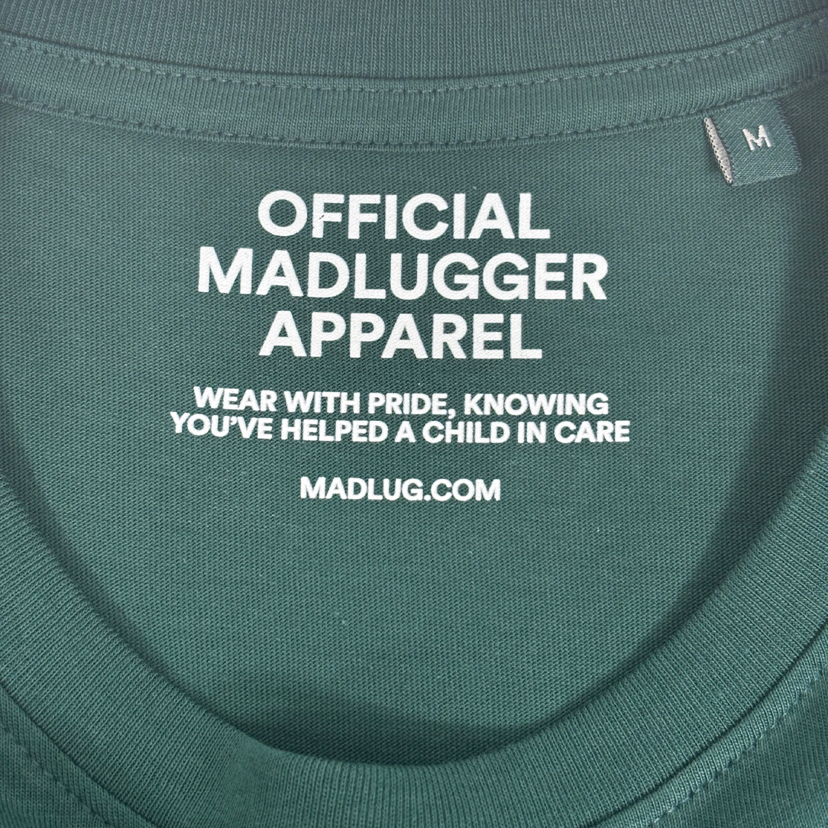 Close-up of a dark green t-shirt neck label with white printed text reading 'OFFICIAL MADLUGGER APPAREL' and 'WEAR WITH PRIDE, KNOWING YOU'VE HELPED A CHILD IN CARE' along with 'MADLUG.COM' 