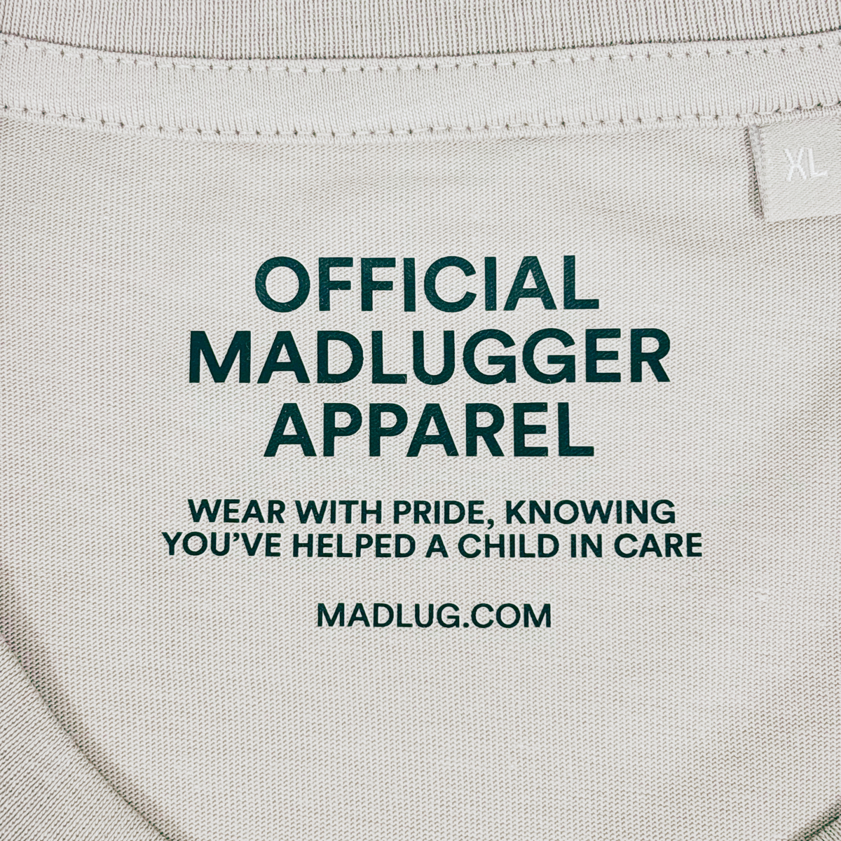 Close-up of a stone colour t-shirt neck label with black printed text reading 'OFFICIAL MADLUGGER APPAREL' and 'WEAR WITH PRIDE, KNOWING YOU'VE HELPED A CHILD IN CARE' along with 'MADLUG.COM' 